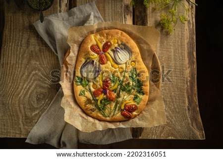Garden art focaccia bread with vegetables, greens, herbs - beautiful flower composition on homemade freshly baked bread, salty vegan pastry. Wooden table. Copy space. Italian cuisine. Top view