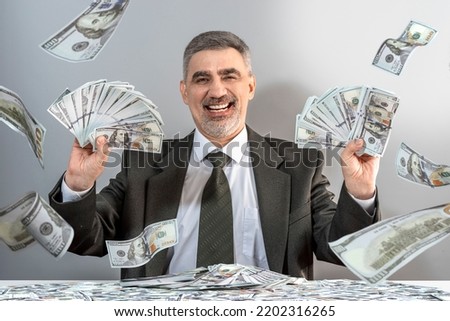 Successful business, income a lot of money, dollar rain concept. Businessman character adult man wearing a formal suit with a tie, and beard, sitting at the table in the office near the gray wall.
