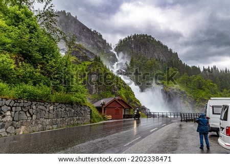 Woman tourist taking pictures of scenic double waterfall Lotefossen. Summer rainy day. The road passes through water jets. Cloud of water dust hangs in the air. Travel to Norway