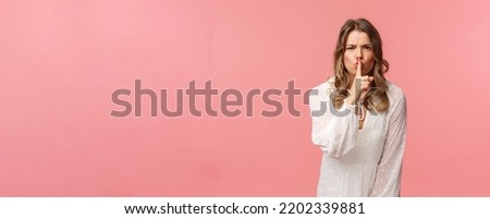 Beauty, fashion and women concept. Angry serious-looking blond girl in white dress, shushing at someone making noise, press finger to lips scolding bad behaviour, say be silent, pink background