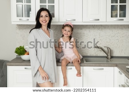 Mother and daughter is smiling and talking at the kitchen. Mom with her little princess at home. Woman and little girl in pink crown together. Facial expression. Selective focus.