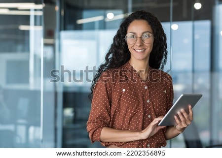Young beautiful and successful business woman with tablet computer and glasses, smiling and looking out the window, Hispanic woman working inside a modern office building at work.