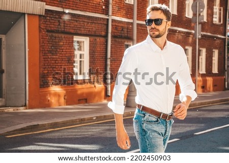Portrait of handsome confident stylish hipster lambersexual model.Modern man dressed in white shirt. Fashion male posing on the street background in sunglasses. Outdoors at sunset. Walking Royalty-Free Stock Photo #2202450903