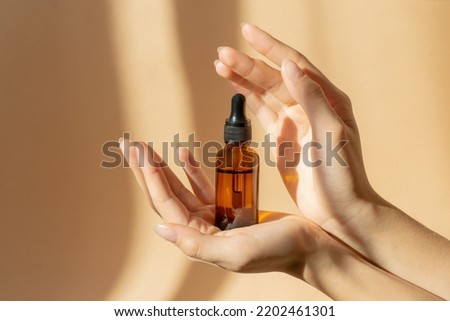Women's hands hold bottle of amber glass with cosmetic serum on brown background. Mockup of container with dropper lid with collagen in rays of sunlight. Concept of skin care Royalty-Free Stock Photo #2202461301