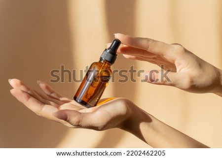 Mockup of glass bottle with dropper lid in female hands. Amber-colored container with cosmetic product, serum on brown background in rays of sunlight. Concept of beauty Royalty-Free Stock Photo #2202462205