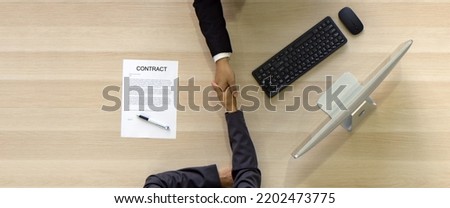 Asian Human Resource Manager in black suit shake hands to congratulate young candidate after the interview. Computer monitor, keyboard, mouse and contract document are on wooden table. Top View