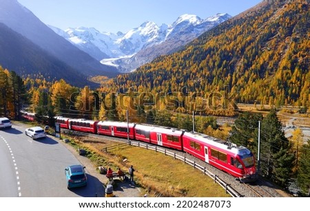 A train travels thru fall colors at Montebello Curve on a brisk autumn day with Morteratsch glacier lying below Piz Bernina and snow-capped alpine mountains in background, in Pontresina, Switzerland Royalty-Free Stock Photo #2202487073