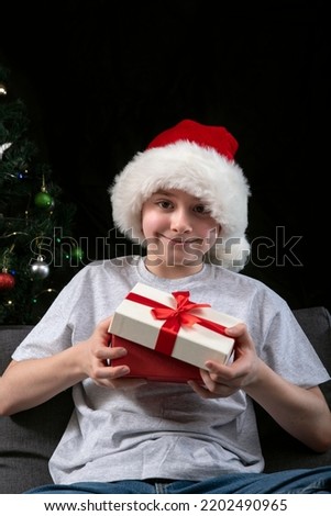 Happy boy in Santa hat opens gift box sitting on the sofa against the background of the Christmas tree. New Year gifts. Vertical frame.