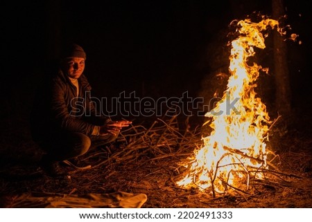 Man sits near flames fire, remnants bonfire with sparks in forest glade at night near riverside. Charred tree branches. burning out firewood. Big bright flame. Close-up background, selective focus
