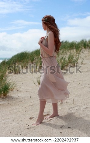full length portrait of beautiful young woman with long hair wearing flowing dress, standing pose walking away from the camera.  ocean beach background with sunset lighting.