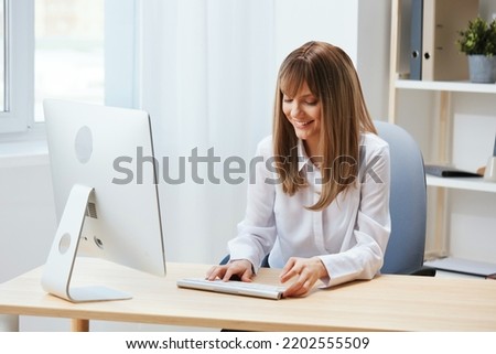 Cheerful happy adorable blonde businesswoman worker freelancer look at screen work on desktop computer online in light modern office. Happy employee writes quarterly report. Copy space, remote job