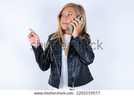 beautiful caucasian blonde little girl wearing biker jacket and glasses over white background speaks on mobile phone spends free time indoors calls to friend.