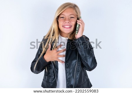 Smiling beautiful caucasian blonde little girl wearing biker jacket and glasses over white background talks via cellphone, enjoys pleasant great conversation. People, technology, communication concept