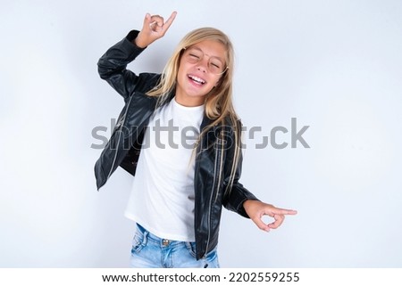 Photo of upbeat beautiful caucasian blonde little girl wearing biker jacket and glasses over white background has fun and dances carefree wear being in perfect mood makes movements. Spends free time o