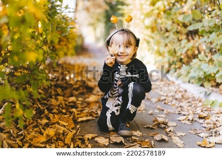 Little toddler boy in halloween skeleton carnival costume sitting on dry leaves in the park in fall. All Saints' day. School autumn hollidays.