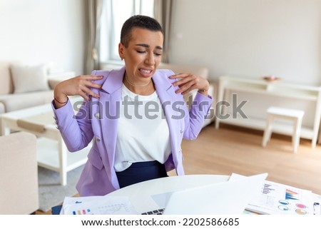 Portrait of young stressed woman sitting at home office desk in front of laptop, touching aching shoulder with pained expression, suffering from shoulder ache after working on pc