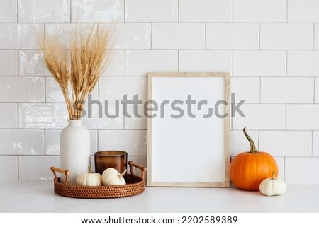 Autumn still life. Nordic kitchen interior with picture frame mockup, tray with vase of dry wheat, candle, pumpkins on white table. Autumn fall, Thanksgiving, harvest concept.