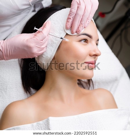Close up of hands of young cosmetologist injecting botox in female face. She is standing and smiling. The woman is closed her eyes with relaxation Royalty-Free Stock Photo #2202592551