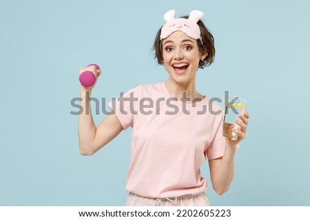 Young excited strong sporty woman in pajamas jam sleep eye mask rest relaxing at home hold dumbbells glass water lemon isolated on pastel blue background studio Healthy lifestyle night bedtime concept