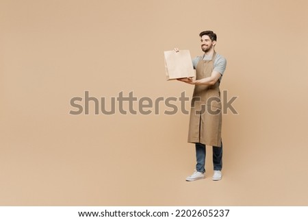 Full body young man barista barman employee wear brown apron work in coffee shop give craft paper bag for takeaway food isolated on plain pastel light beige background. Small business startup concept Royalty-Free Stock Photo #2202605237