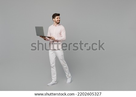 Full body young caucasian IT man 20s he wearing basic white shirt hold use work on laptop pc computer walk go look aside on workspace area isolated on plain grey background. People lifestyle concept