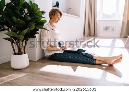 Side view of focused barefoot businesswoman sitting with crossed legs on floor near potted green plants and working on remote project while using computer