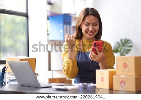 Woman in a small start-up business woman working in a home office working on a laptop and smartphone checking orders from the internet in preparation for shipping. Royalty-Free Stock Photo #2202619041