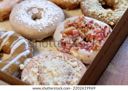 Picture of assorted donuts in a box with chocolate frosted, powdered and bacon sprinkled donuts.