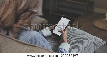 close-up image, Beautiful young Asian female using her smartphone while relaxing in her minimal living room. a woman holding a mobile phone white screen mockup.
