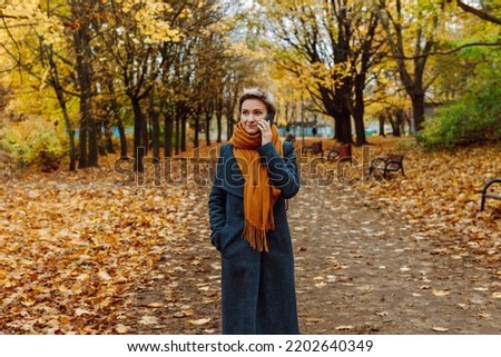 A blonde in a coat and scarf talks on the phone as she walks through an autumn park Royalty-Free Stock Photo #2202640349