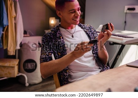Young handsome stylish smiling boy looking at polaroid pictures, while sitting by table in cozy room at home