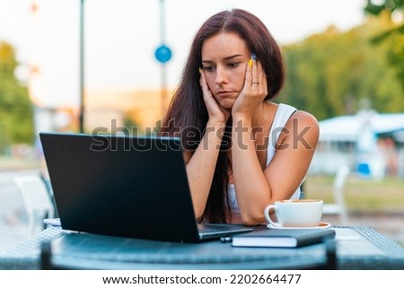 Tired or sad,exhausted beautiful woman freelancer working on her laptop in cafe outdoors,holding her head with hands, looking at screen.Overworked,deadline,bad news concept.Summer day.