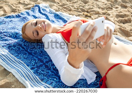 Beautiful young tourist woman on holiday relaxing on a beach and holding a smartphone device in her hands taking selfies pictures of herself, networking. Travel vacation technology outdoors.