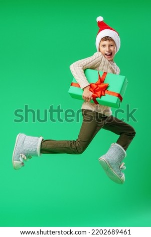 Happy smiling boy with a gift box with a bow in his hands and a red Santa hat on his head jumps on a green background. Preparation for Christmas, sale at a discount. Dynamic image.