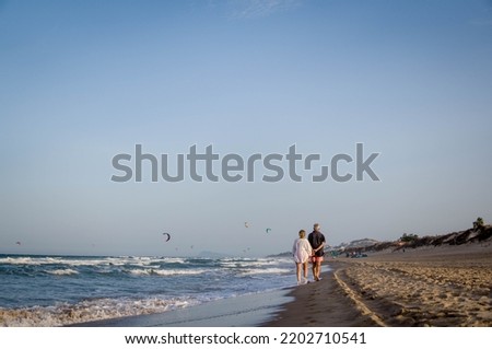 Silhouette of Elderly Couple Walking Barefoot Calmly at Sunset along the Shore of the Beach with Kite Surfers in the Background in the Sky