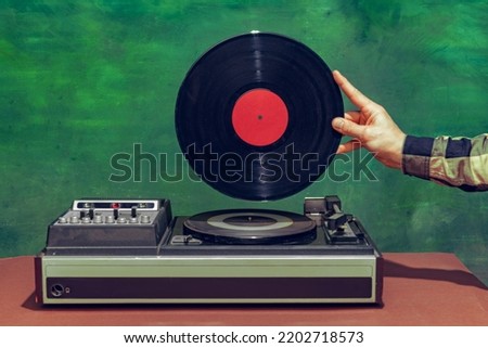 Colorful image of male hand holding retro vinyl record in front of vintage player isolated over dark green background. Concept of pop art, fashion, music, mix old and modernity. Copy space for ad Royalty-Free Stock Photo #2202718573