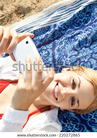 Over head portrait of a young tourist woman on holiday relaxing on a beach and holding a smartphone device in her hands taking selfies pictures of herself. Travel vacation technology outdoors.