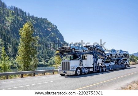 Industrial Big rig white classic car hauler semi truck tractor transporting cars on the modular hydraulic two level semi trailer driving on the one way highway road along the river in Columbia Gorge Royalty-Free Stock Photo #2202753961