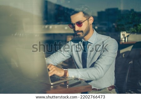 Portrait of handsome business man wearing sunglasses working on laptop in modern office. Through glass view.