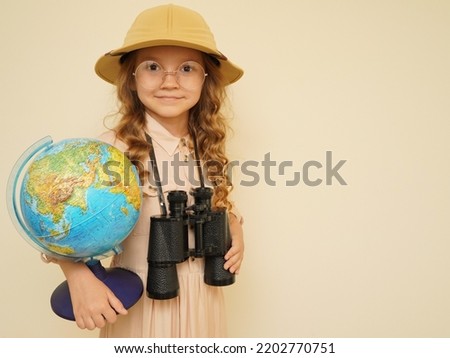 Happy child plays the discoverer. Unique child. Travel, adventure, vacation and discovery concept. A kid in a traveler's hat with binoculars and a globe in his hands on the beige background.  Royalty-Free Stock Photo #2202770751