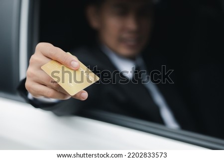 An Asian man in a formal suit stays in the car, opens the window, handed his credit card to pay for gas, travel by car, safe driving, respecting traffic rules. Royalty-Free Stock Photo #2202833573
