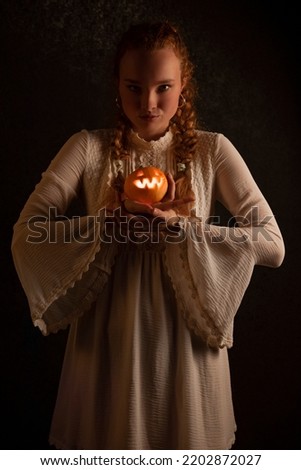 Woman stands and holds a small Jack's pumpkin lamp in her hands on a black dark background in a white dress. Concept of halloween.