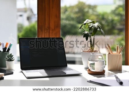 Laptop with programming code on computer screen, stationery and potted plant on white table