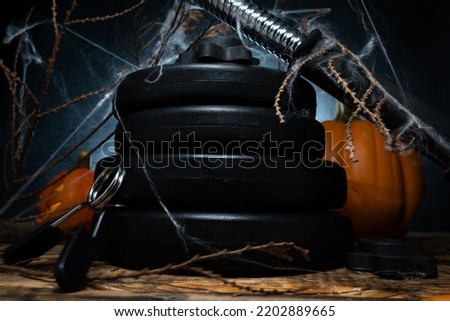Heavy gym dumbbell barbell weight plates stacked on top of each other, covered with spider web. Healthy fitness lifestyle autumn or fall composition for Halloween, with pumpkins in background.