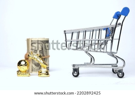 Selective focus picture of shopping cart, golden ball and boot trophy miniature with fake money insight. Football player transfer window concept.