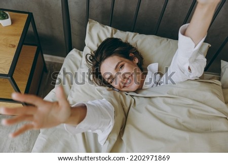 Young woman wears casual white shirt she lying in bed rest relax spend time in bedroom lounge home in own room hotel wake up stretch hands dream be lost in reverie good mood day. Lazy morning concept Royalty-Free Stock Photo #2202971869