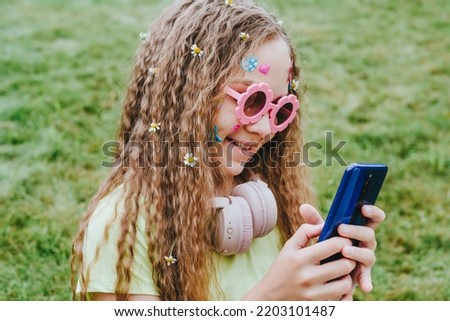 Cute teenage girl with stickers on her face wearing pink glasses and headphones taking selfie on smartphone with chamomile flowers in curly hair posing on green grass background.