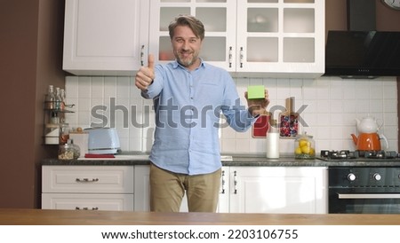 A young man is holding something green in the kitchen, showing a product, smiling and presenting a cheerful, imaginary object. 