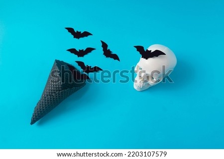 A black ice cream cone with bats flying out and a white scull with a bat on the forehand. Blue background. Minimal surreal Halloween creative concept for banner or advertisement