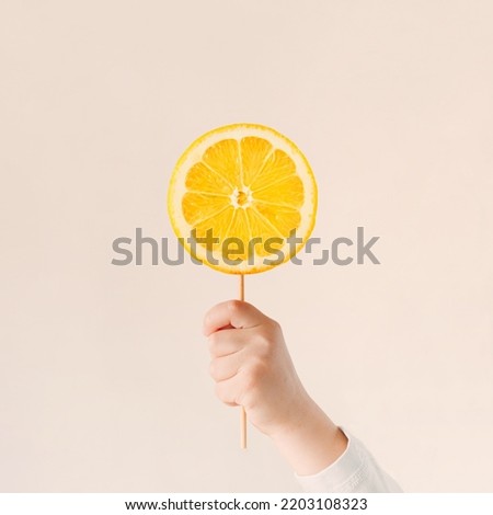 Child’s hand holding stick with lemon slice on pastel beige background. Creative candy scene. Natural organic vitamins idea. Raw citrus fruit lollipop. Healthy sweets concept. Minimal food design. Royalty-Free Stock Photo #2203108323